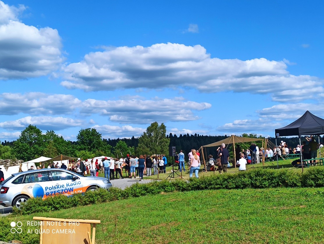 Krajobraz z ludźmi, drzewami i białym samochodem z napisem "Polskie Radio Rzeszów" w słoneczny dzień z chmurami na niebie.